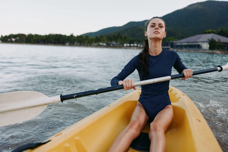 Athletic woman rows a yellow kayak across calm water, gripping a paddle with effort while ripples trail behind. Distant shoreline and mountains frame the scene.