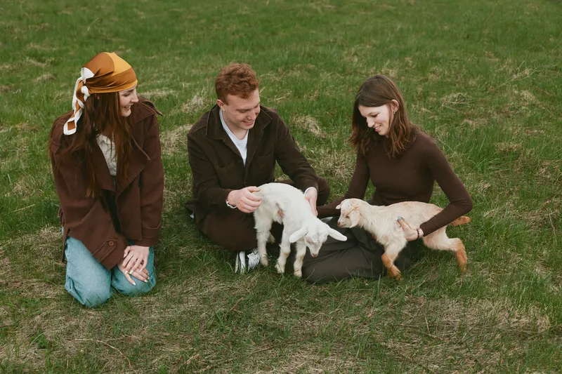 Three people enjoy a playful moment outdoors as goats interact with friendly kids, friends smile and pet the animals on green grass, showing casual countryside lifestyle and warm bonding in a candid