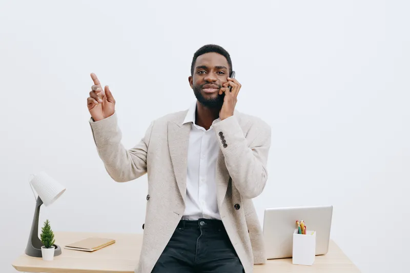 Confident African American man in smart casual attire talking on mobile phone while gesturing with his hand in modern office setting isolated on plain white background. Business communication concept,