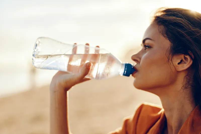 Woman drinking water from plastic bottle in bright outdoors, side profile, focused on hydration and thirst relief in summer heat, healthy lifestyle concept for wellness and routine care