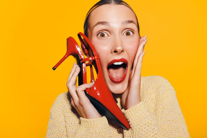 Excited woman with red lipstick holds red high heel shoes near face on bright yellow background. Concept of fashion, style, shopping, and happiness with vibrant studio lighting.