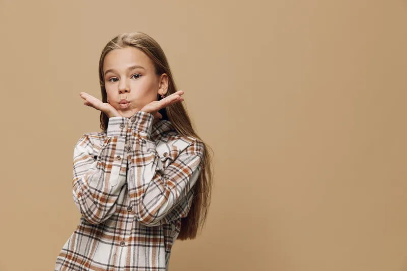 Girl with long hair wearing checkered shirt posing with hands near face on beige background. Child with playful expression and casual clothes in studio portrait with neutral backdrop.