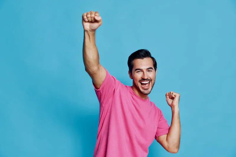 Happy cheerful young man celebrating with hands raised, wearing a pink t-shirt, smiling broadly against a solid blue background. Joyful lifestyle concept.