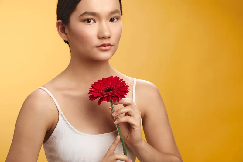 asian woman holds a red flower against a warm yellow backdrop, presenting a calm, thoughtful portrait with soft lighting and a clean, minimal composition.
