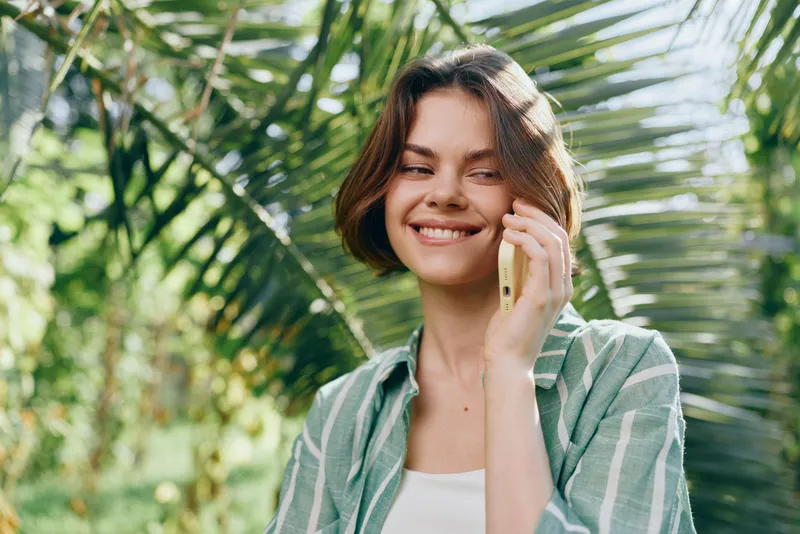 Portrait of a cheerful woman talking on a smartphone outdoors, surrounded by lush tropical plants.