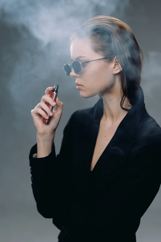 Young woman wearing sunglasses vaping with smoke around her in a dark atmosphere