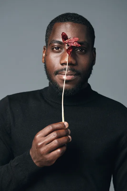 African man with dark skin holding dried flower in front of face in creative conceptual visual style. Portrait with black turtleneck and neutral background for artistic expression and culture.