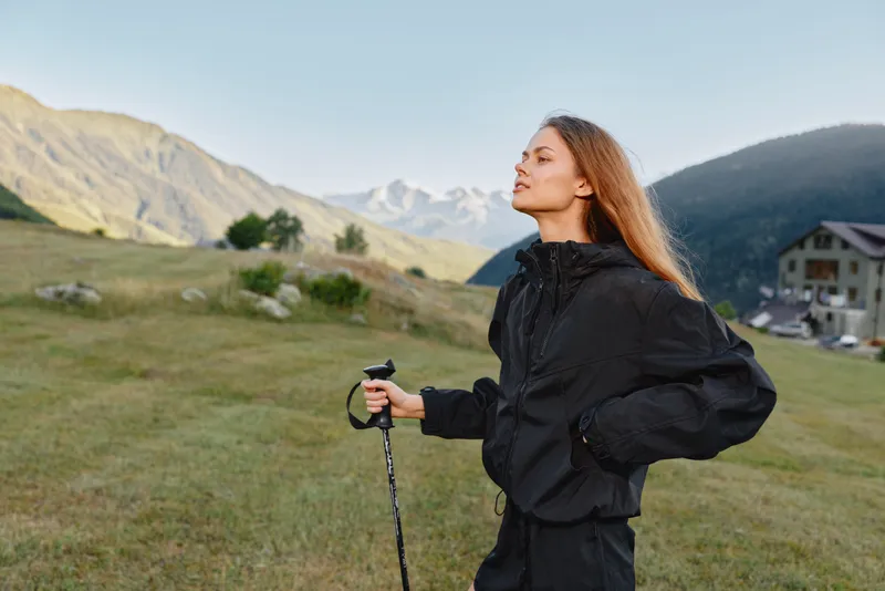 A woman wearing a black jacket stands in a grassy field with a trekking pole, looking toward distant mountains. The open landscape exudes calm, adventure, and outdoor freedom.