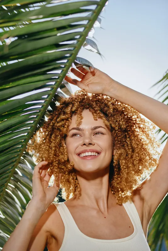 smiling woman with curly hair enjoys sunny tropical outdoors surrounded by green palm leaves and clear blue sky, radiating happiness and natural beauty.