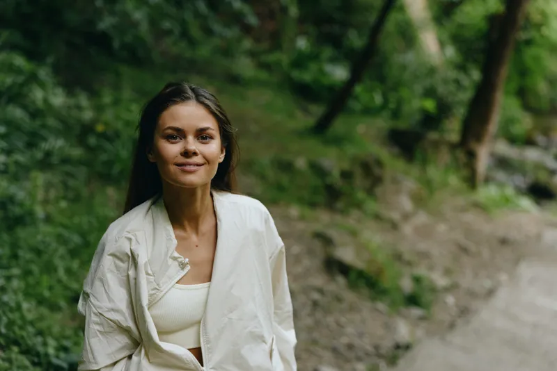 smiling woman outdoors wearing white jacket stands on woodland path surrounded by green trees, natural light highlighting her relaxed and peaceful expression in spring.