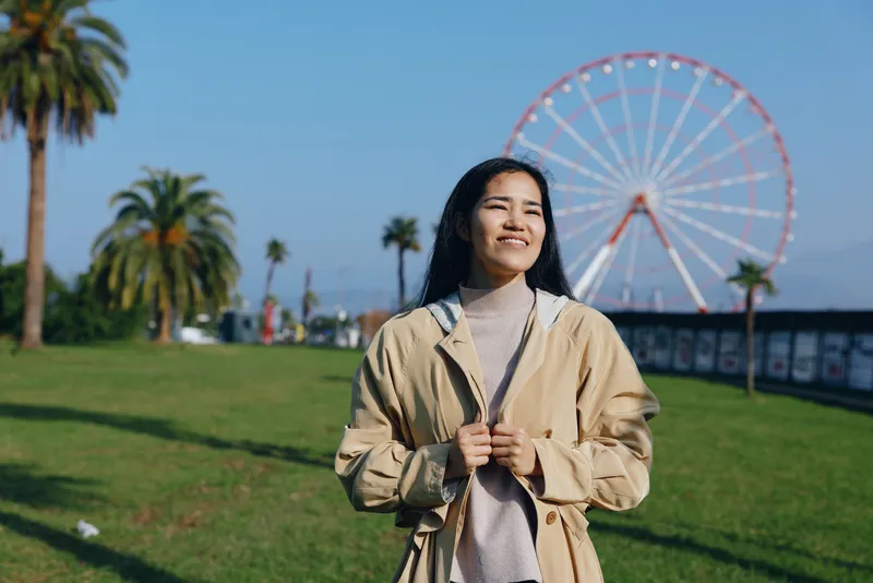 Woman in a light tan jacket standing in a sunny park with a large ferris wheel in the background, casual pose, cheerful mood, relaxed fashion and blue sky behind
