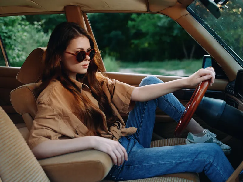 A young woman with long brown hair, wearing oversized sunglasses and a stylish shirt, relaxing in a vintage car, embodying a sense of freedom and adventure on a sunny day.