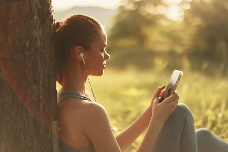 Young woman enjoying music outdoors in nature with smartphone and earphones, wearing sportswear, conveying relaxation and joy in a sunny setting