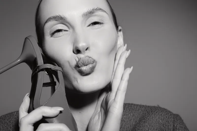 Young white woman with fair skin and no makeup holds a red high heel shoe close to her face, puckering lips in a playful pose in studio lighting.