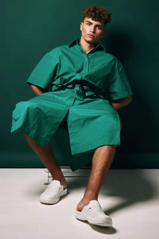 Young man sitting with hands behind back wearing oversized green shirt dress and white slip-on shoes against solid green background in studio photo shoot pose.