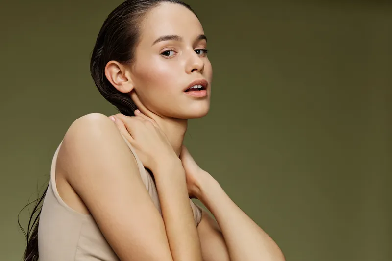 Fashion portrait of a young model with long dark hair, wearing a beige top, showcasing natural beauty against a soft green background