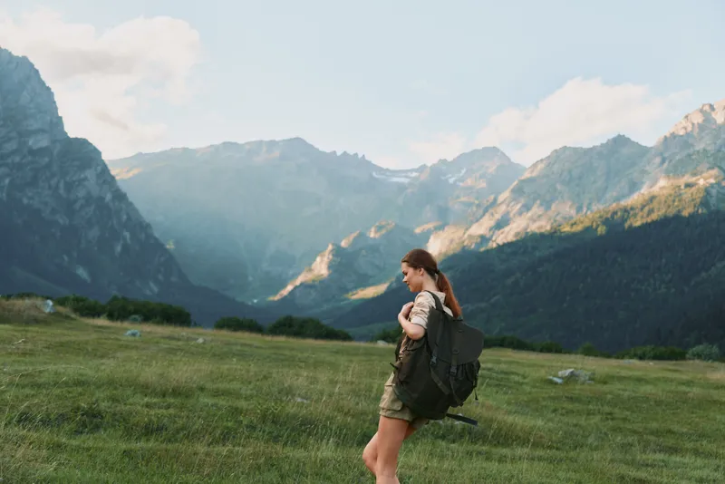 Hiker with a large backpack strides across a sunlit alpine meadow under towering mountains, embodying freedom and adventure in a single warm moment