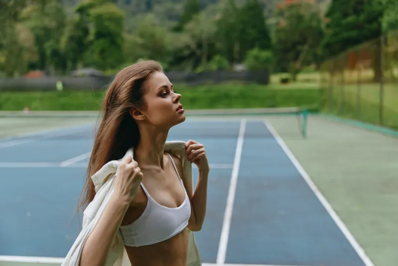 Young athletic woman in sportswear confidently posing on a tennis court, showcasing healthy lifestyle and fitness, seasonal summer sports concept, vibrant greenery