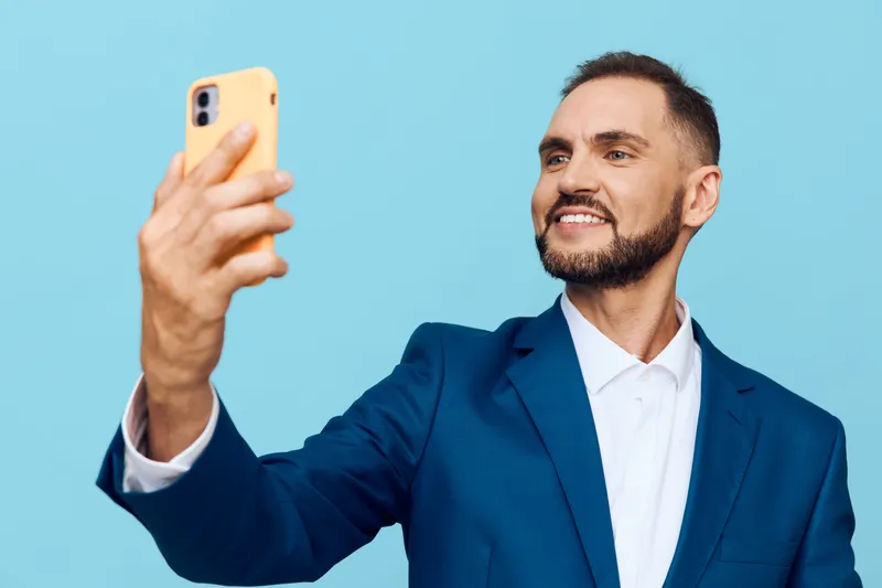A confident business professional man smiles while taking a selfie against a solid colored background in a studio. The image conveys pride, joy, determination, and forward facing energy.