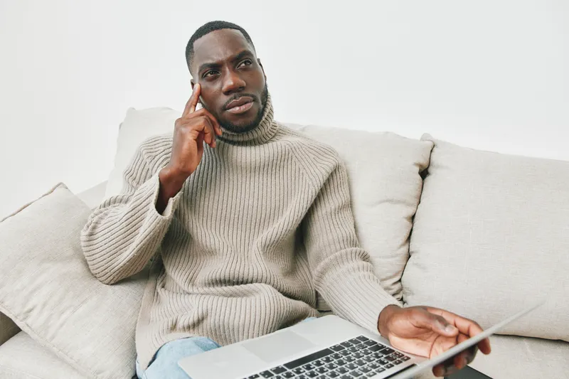 Thoughtful man in a cozy sweater seated on a couch, talking on the phone while using a laptop, reflecting a modern lifestyle and remote work concept