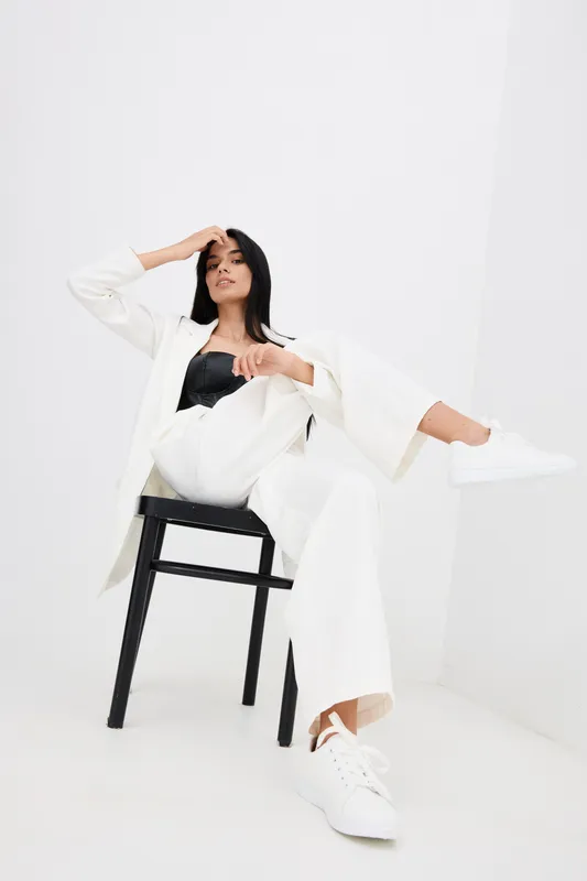 stylish woman wearing white pantsuit and sneakers sits relaxed on black chair in minimalistic bright studio, showcasing modern fashion and casual elegance