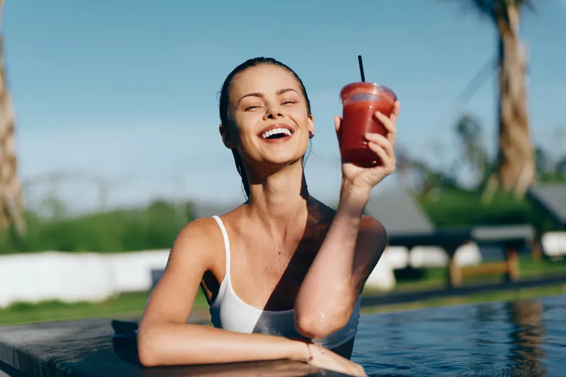 Smiling woman in a white swimsuit rests at poolside, raising a red smoothie. Bright outdoor setting evokes wellness, hydration, leisure, and carefree mood with warm sun, clear blue water, and relaxed