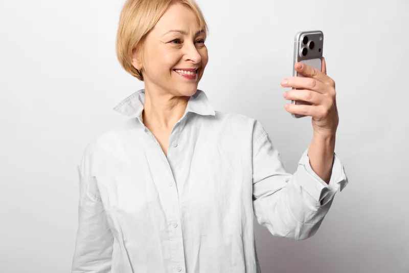 Woman with blonde short hair shows smile while holding smartphone for a selfie, portrait on white studio background, casual mood, modern technology and communication now