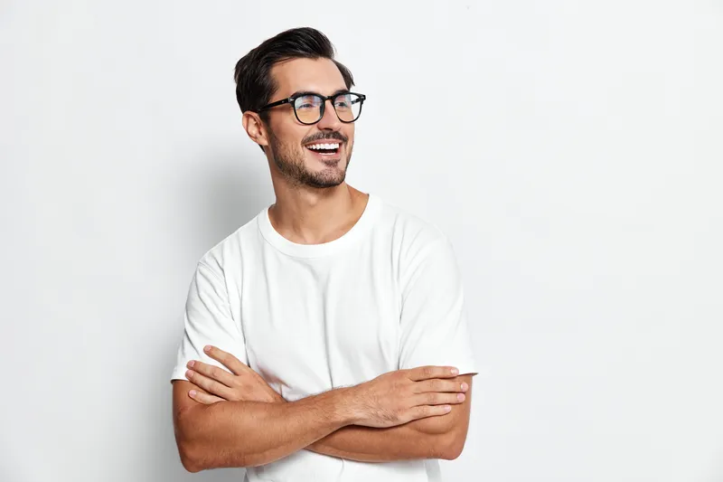 Cheerful smiling man wearing glasses and white t-shirt, looking away with a joyful expression. Isolated on neutral gray background, showcasing happiness and confidence.