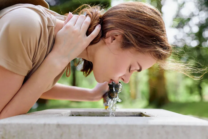 Woman drinking from a park fountain outdoors, closeup portrait sideview of young adult female with brown hair hydrating on a sunny summer day, candid leisure and refreshment.