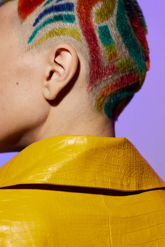 Close-up of a woman with colorful patterned hair and a yellow textured jacket against a purple background, showcasing creative style and bold fashion choices.