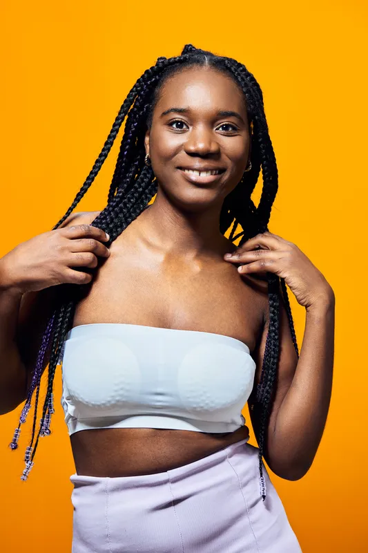 Young Black woman with braids smiling confidently against a bright yellow background, wearing a light top and skirt, exuding joy and positivity in a vibrant and trendy style
