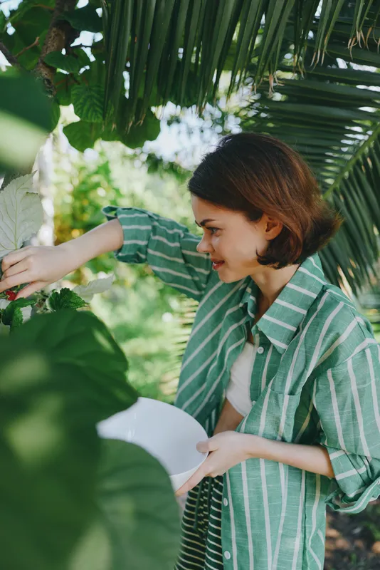 Woman examining lush tropical foliage outdoors, wearing a green striped shirt, with a white plate in hand amid vibrant leaves.