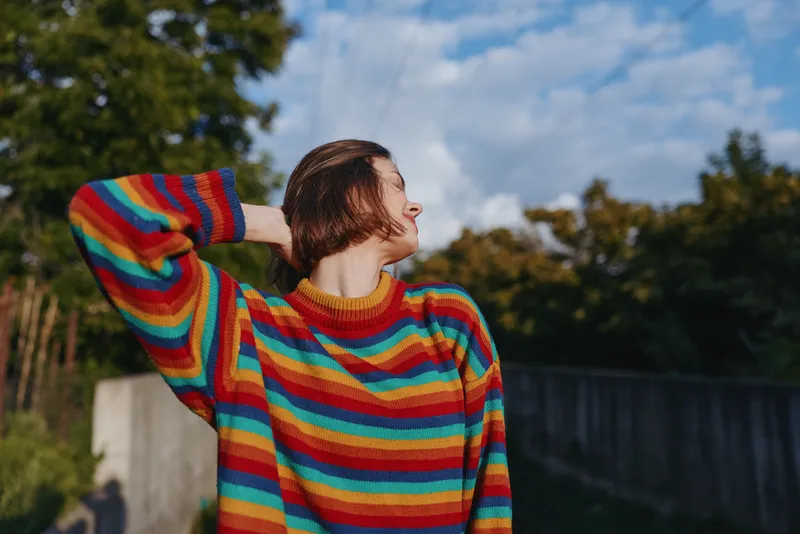 Woman, young woman, striped sweater, short hair, smiling, outdoor, casual outfit, sunshine — A 20s model in a colorful rainbow knit sweater smiling and leaning back with hand behind head in sunny