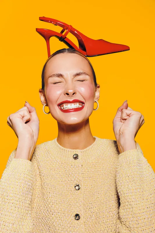 Happy woman with closed eyes smiles broadly wearing beige sweater and red high heel shoe on head. Bright orange background creates cheerful and playful mood in studio portrait.