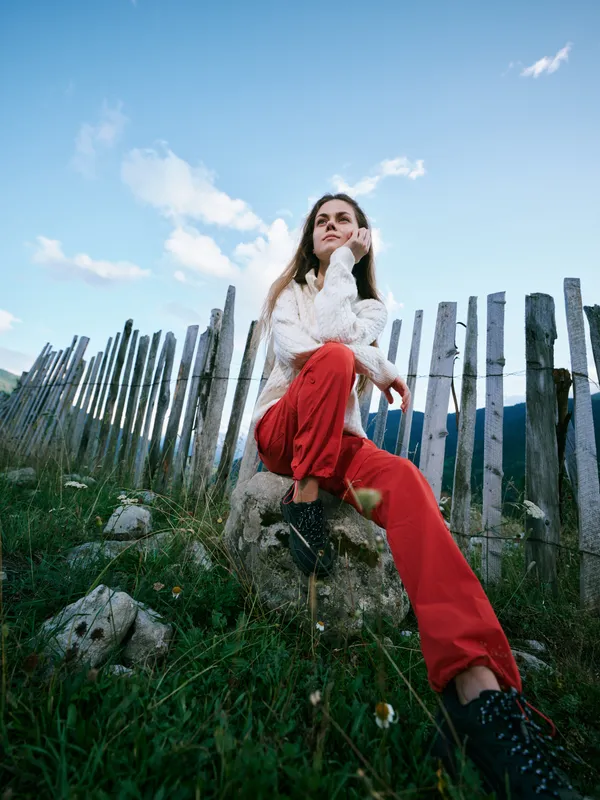 Female figure sits on a rock beside a weathered fence, red pants and boots, serene hillside landscape under a blue sky with clouds, casual outdoor styling and relaxed mood.