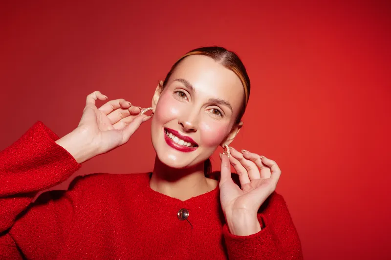 Smiling woman in red sweater adjusts earrings on red background. Close-up portrait with bright makeup and slicked back hair. Concept of fashion, beauty, and confident style.