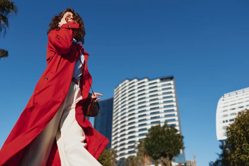 A woman with curly hair wearing a red coat strides through a sunny urban street, with tall modern buildings behind her, conveying confidence, motion, and bold street style.