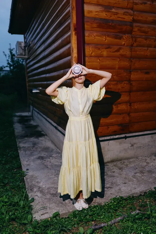 woman portrait in yellow dress by wooden house holding cup to eyes like binoculars outdoors summer scene authenticity inclusivity playful moment