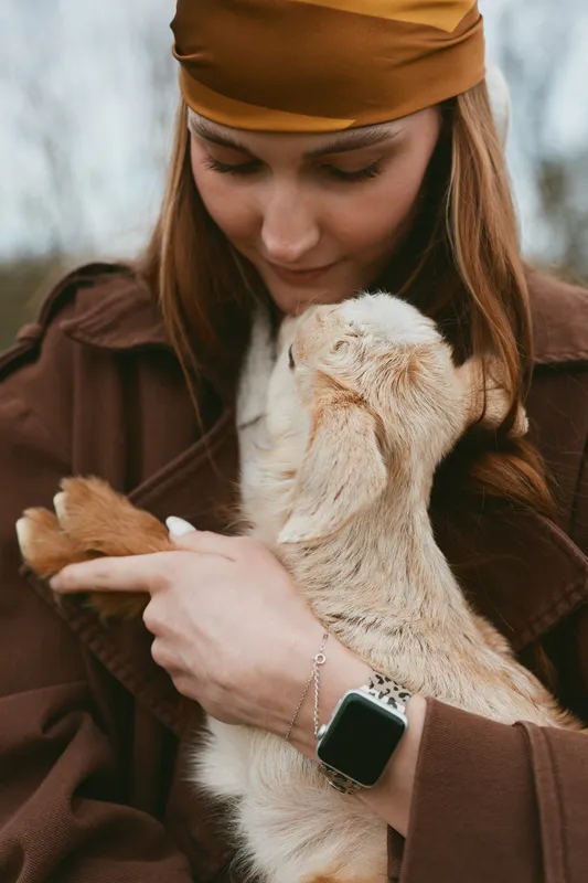 Lifestyle moment with a woman in a brown coat and headband gently holding a goat kid, capturing bonding, care, and cozy outdoor autumn mood.