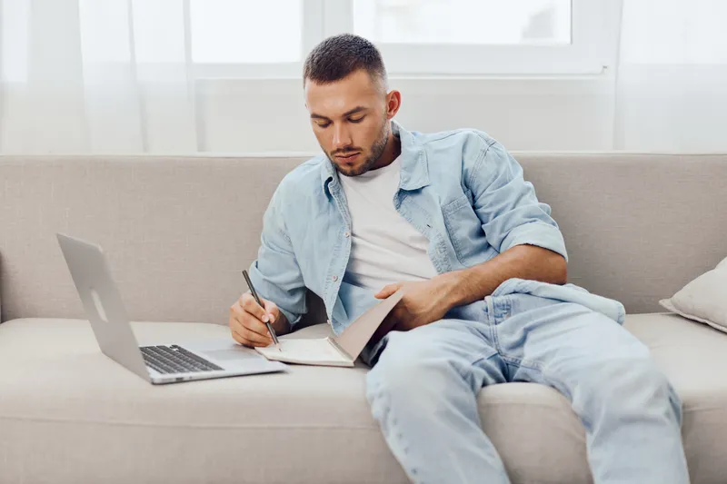 Thoughtful man reflecting on his work-life balance and mental health while enjoying a digital detox, seated on a cozy couch with a laptop nearby.