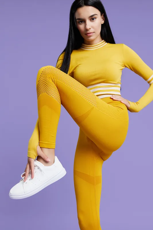 Sport fitness woman in bright yellow activewear poses with one leg raised against purple background, showing focus, strength, and healthy lifestyle in vibrant athletic style.