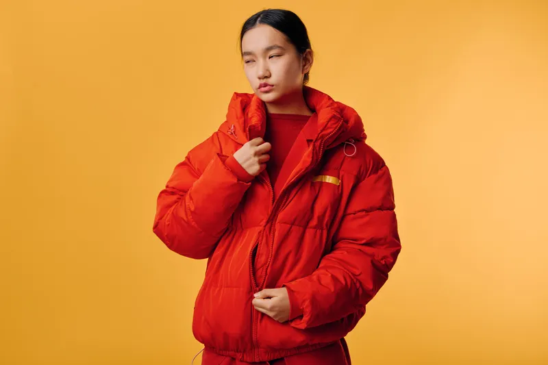 Young woman wearing warm red puffer jacket and red sweater posing against yellow studio background with eyes closed, fashion portrait for winter style and seasonal clothing concept.