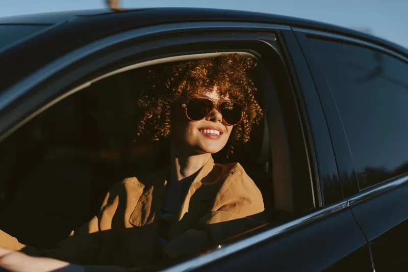 Smiling woman with curly hair in a car wearing sunglasses. Warm retro film color emphasizes sunny mood and casual lifestyle, capturing a moment of everyday freedom and relaxed confidence.