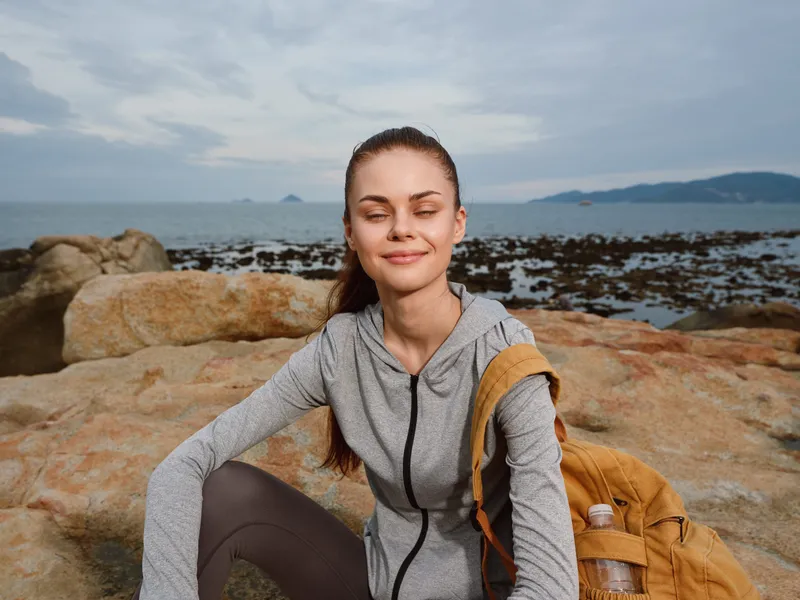 Smiling young woman in sporty outfit sitting on rocks near the sea, enjoying nature and a healthy lifestyle, with a backpack and tranquil background