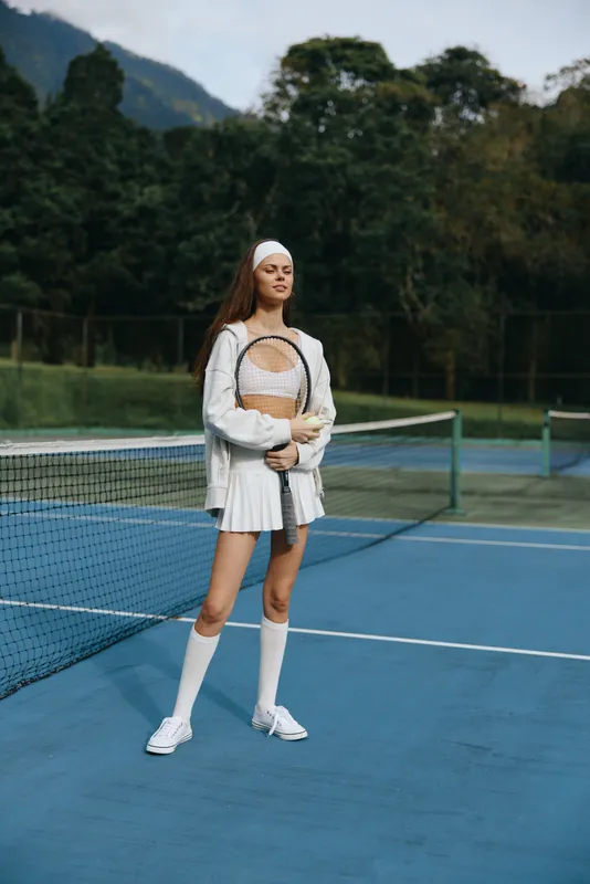 Athletic young woman in sporty tennis attire, exuding confidence and joy, ready to play on a vibrant blue tennis court surrounded by nature