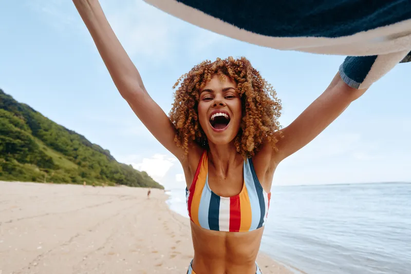 Young woman with curly hair joyfully raises her arms while standing on a sandy beach wearing a colorful striped swimsuit under a bright sky.