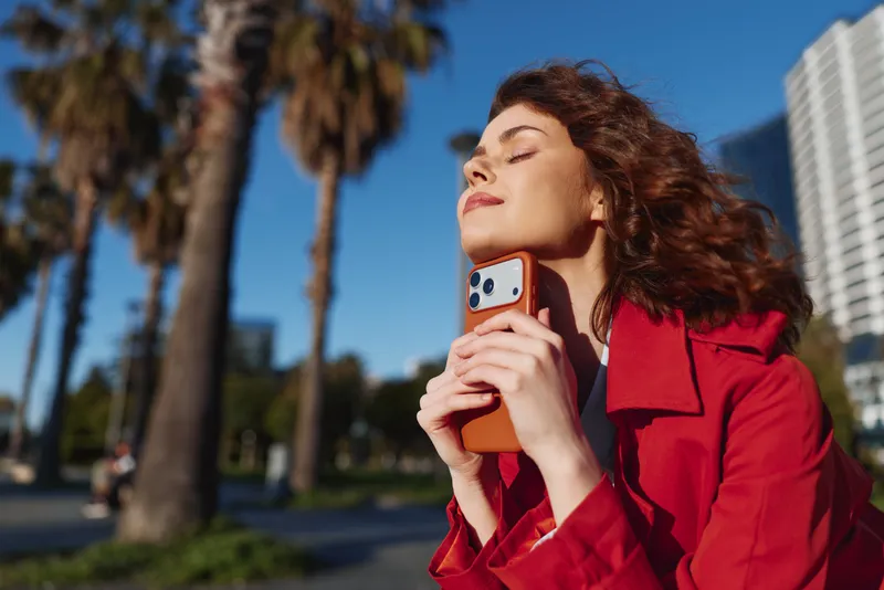 Cheerful woman outdoors, sunlit moment with closed eyes, red coat blown by breeze, holding phone, lush greenery and palm trees in background, natural colors, relaxed mood, vibrant urban park scene
