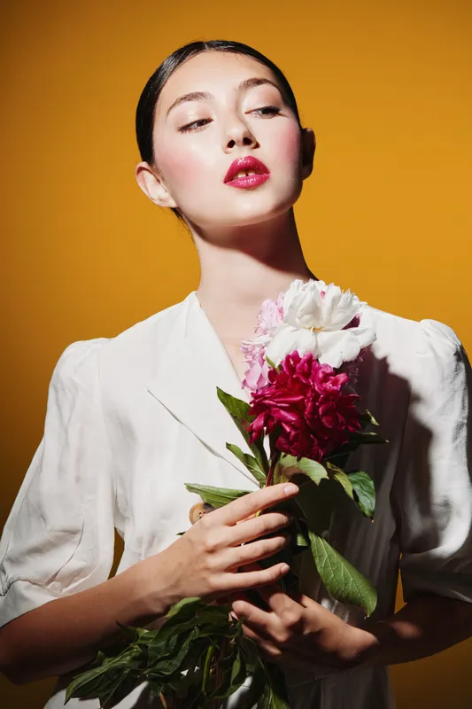 Fashion creative portrait of a young woman with sleek hair and glossy red lips holding flowers. Clean white outfit contrasts with vibrant yellow background. Beauty concept.