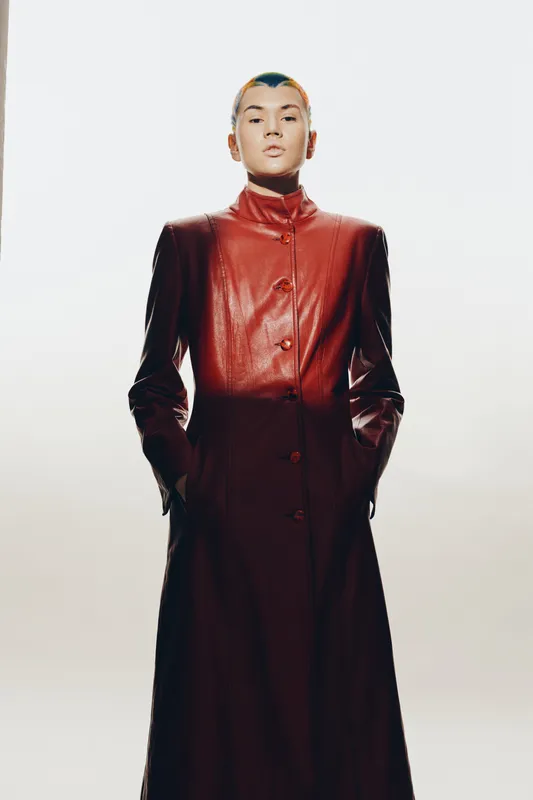 woman in long red leather coat standing with hands in pockets, minimalist background, short hair, serious expression, fashion attitude
