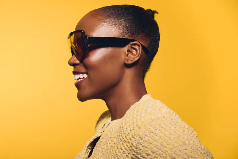 Profile of a smiling black woman with dark skin wearing large sunglasses and a yellow knitted top. Bright yellow background creates a cheerful and stylish mood with natural beauty concept.